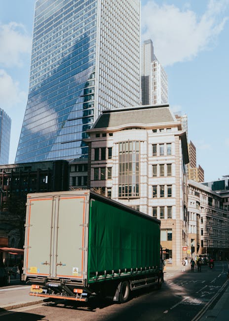 A white moving van with a closed cargo area is parked on a city street in front of a large historic building with elaborate stone architecture and multiple arched windows. To the left, there are sever